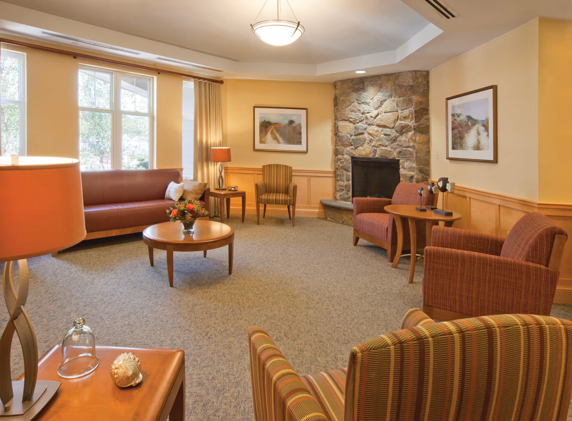 Well-lit senior living common room with sofas and armchairs arranged around a coffee table and a stone fireplace.
