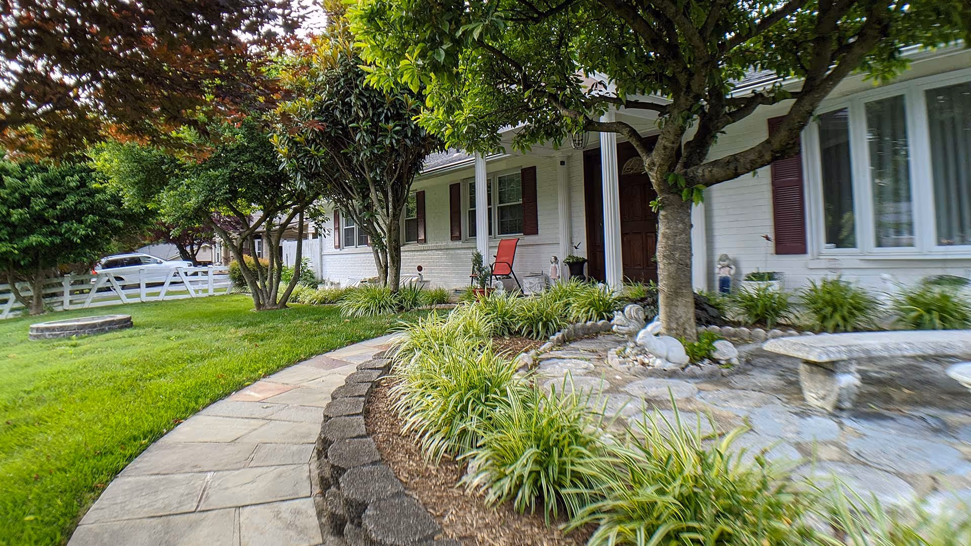 A landscaped front yard of a white brick house with a curved stone pathway, green grass, various trees and plants, a stone bench, and garden decorations including a white rabbit statue. A red chair is placed near the front door under a covered porch.