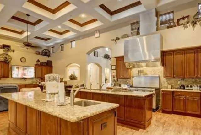 Spacious kitchen area with two large granite countertops, wooden cabinets, a stainless steel stove with a large hood, and a sink. The ceiling features recessed lighting and decorative beams. In the background, there is a living area with a mounted TV and decorative items on shelves.