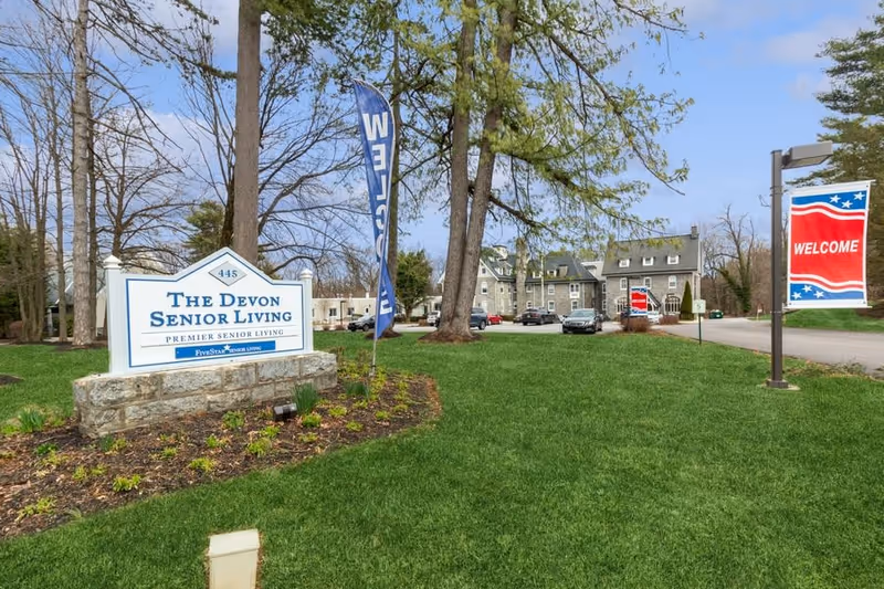 Outdoor view of The Devon Senior Living facility with a large sign displaying the facility name and a well-maintained lawn. There are trees, a parking area with several cars, and banners that say 'WELCOME'. The building is visible in the background under a partly cloudy sky.