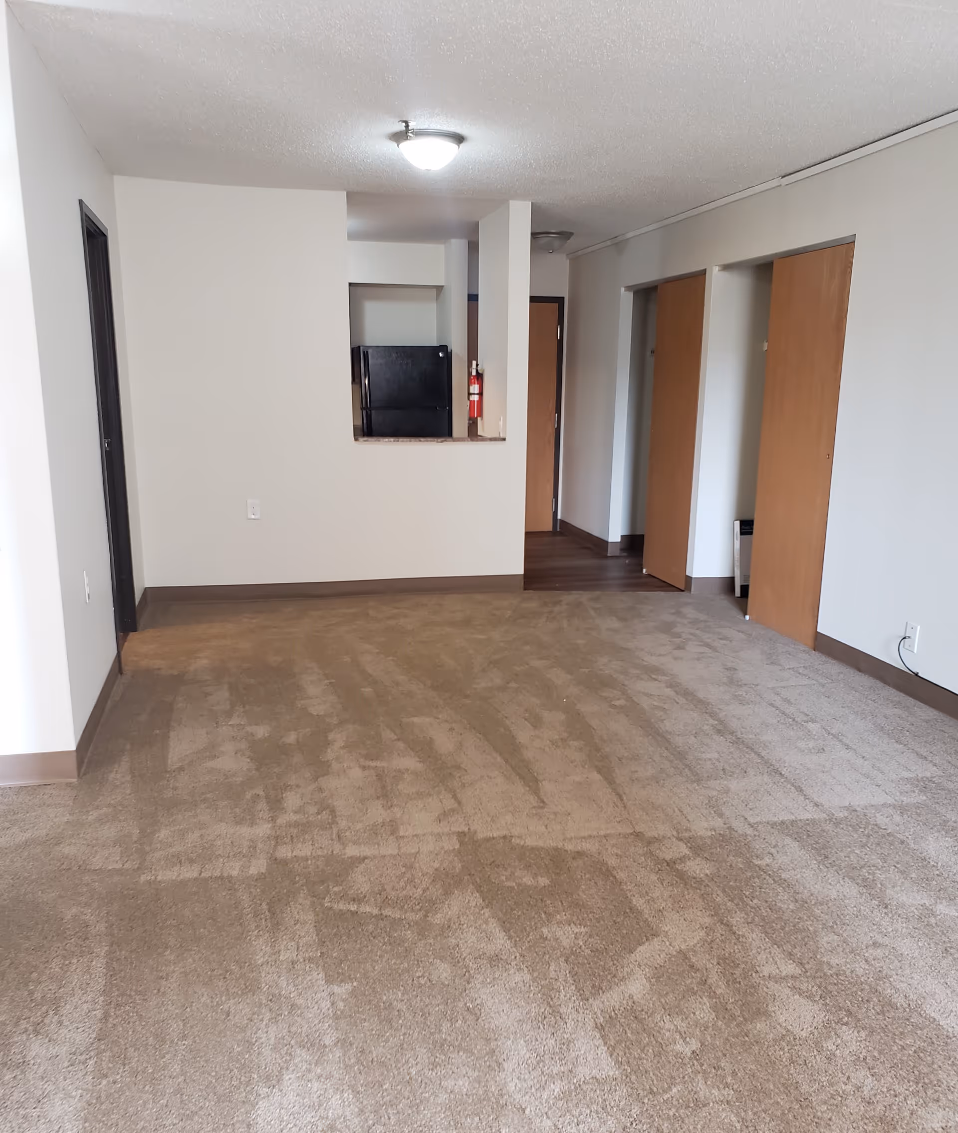Empty apartment room with beige carpet, white walls, and a ceiling light. There is a small kitchen area with a black refrigerator visible through an open wall space. Two wooden doors are on the right side, and a hallway with a wooden floor leads to another door in the background.