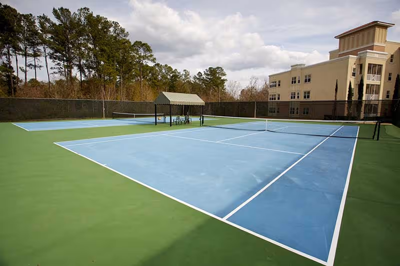 Outdoor tennis courts with blue playing surfaces and green surrounding areas, a small shaded seating area with chairs in the center, trees in the background, and a multi-story building to the right under a cloudy sky.