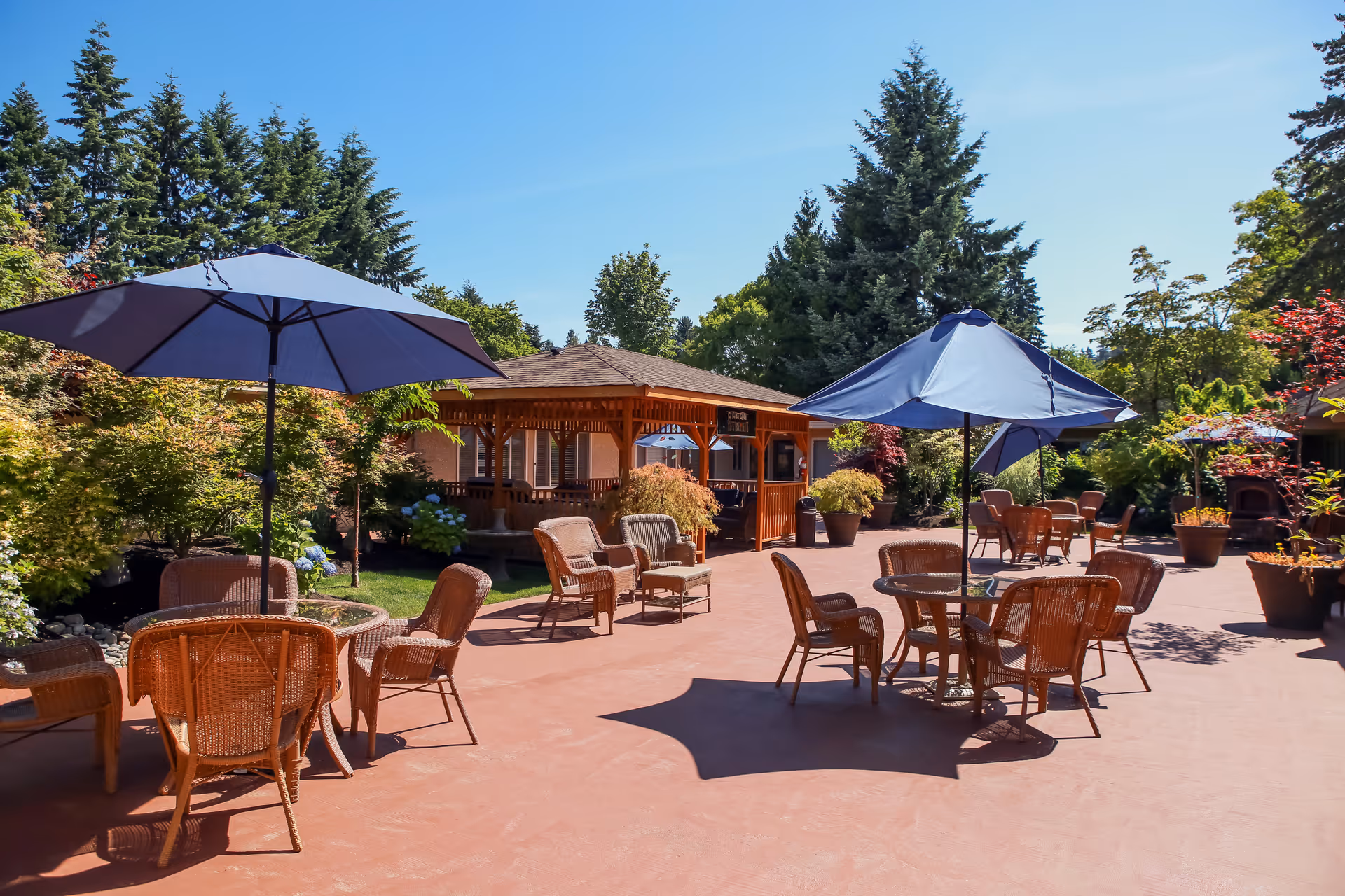 Outdoor patio area with several round tables and wicker chairs, each table shaded by large blue umbrellas. The patio is surrounded by lush greenery, potted plants, and tall trees under a clear blue sky. A wooden gazebo structure is visible in the background.