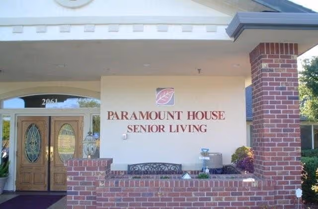 Entrance of Paramount House Senior Living facility featuring double wooden doors with decorative glass panels, a brick wall with a black metal bench, and the facility name displayed on the wall above the bench.