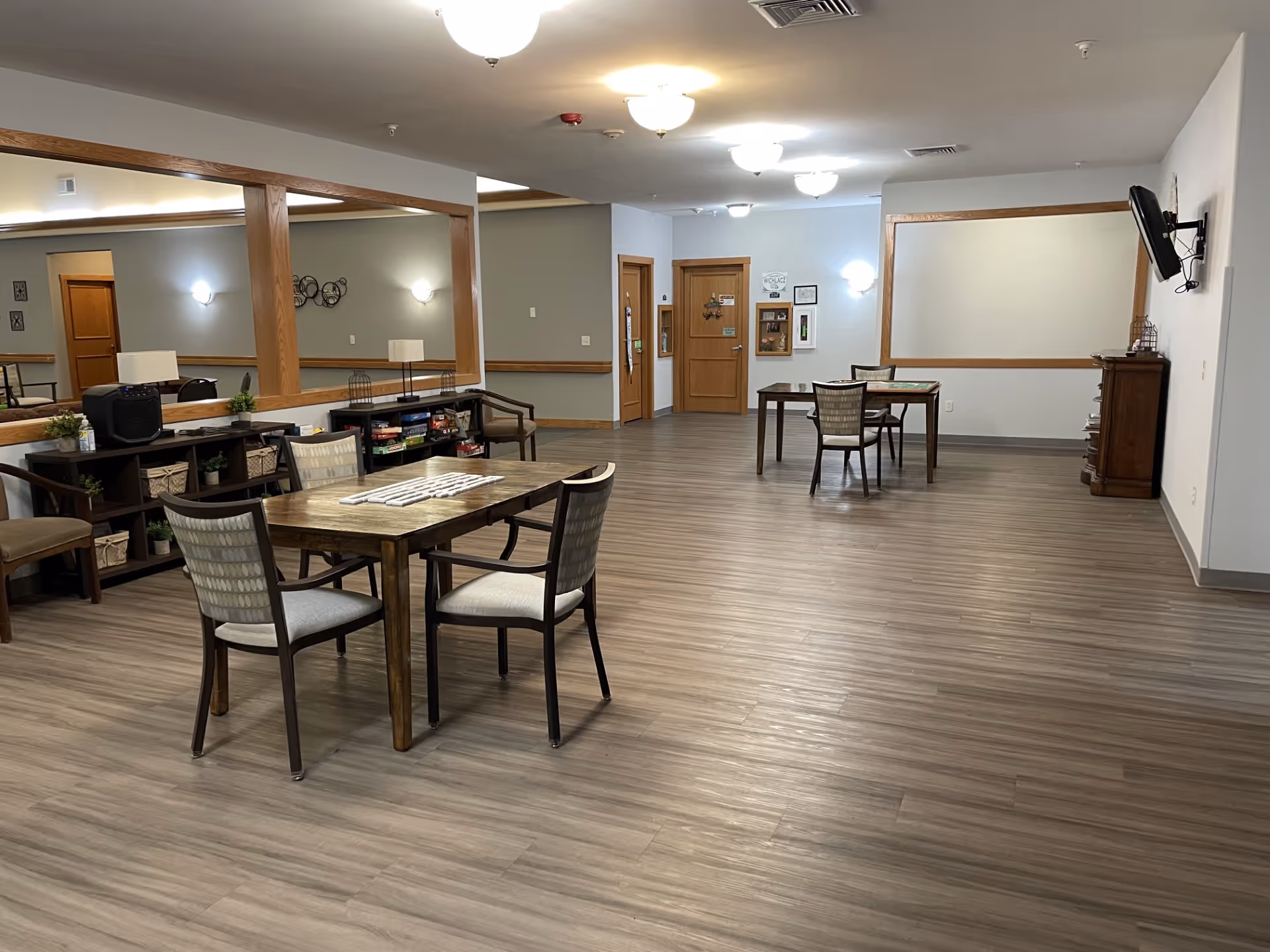 Spacious communal activity room with several tables and chairs, shelving with games and decor, and wood-look flooring.