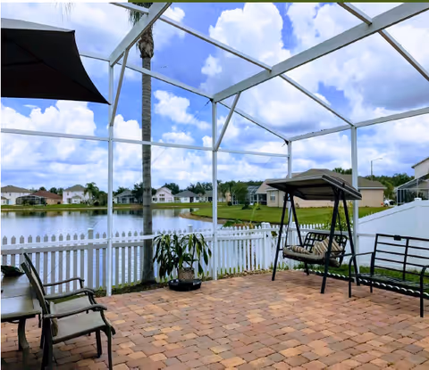 A screened-in patio area with brick flooring, outdoor chairs, a swing bench, and a small table. The patio overlooks a white picket fence, a palm tree, a pond, and houses in the background under a partly cloudy sky.