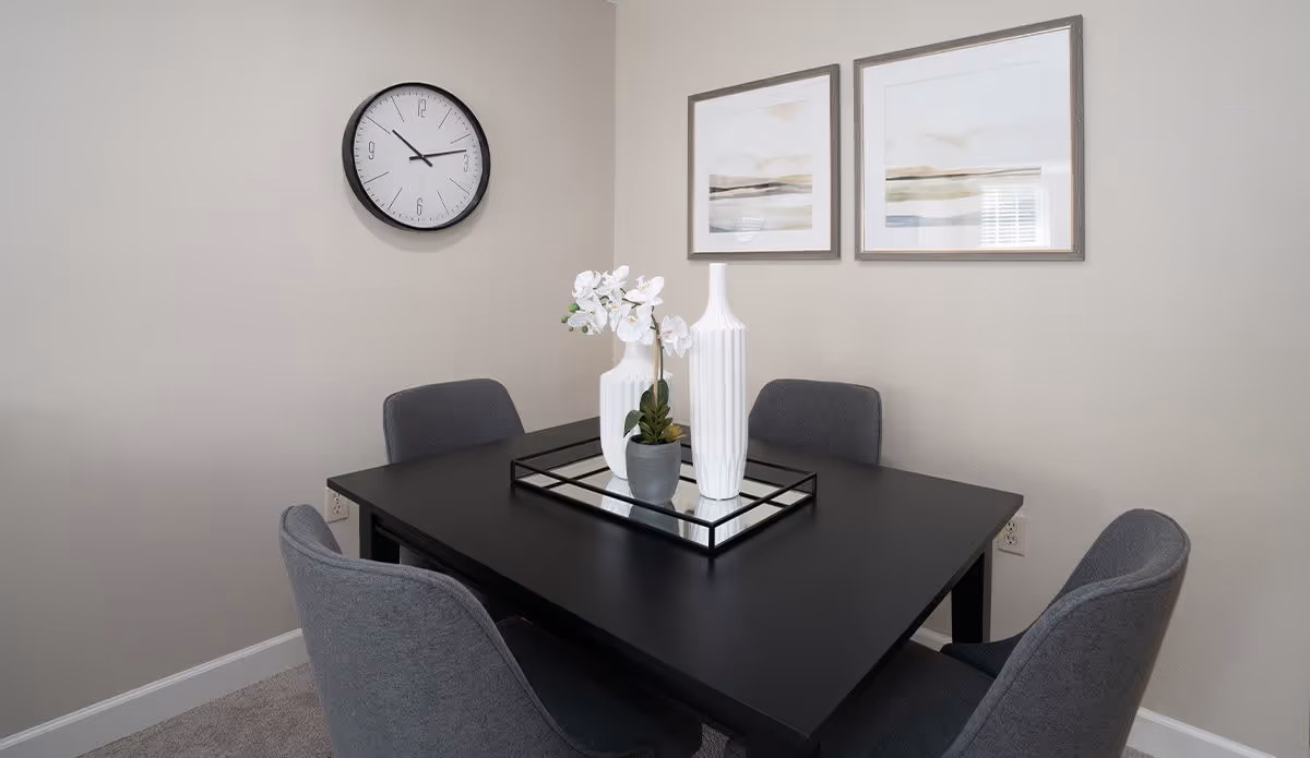 Small dining area with a black table surrounded by four gray chairs, decorative vases on a mirrored tray, a wall clock, and framed artwork on the walls.