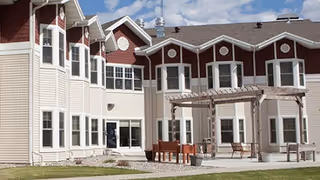 Two-story building with beige siding and red upper facade, multiple bay windows, a pergola and outdoor seating on a lawn.