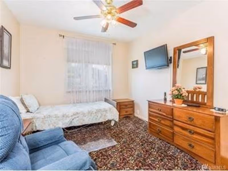A cozy bedroom with a single bed covered in a floral bedspread, a blue upholstered armchair, a wooden dresser with a mirror and a potted plant, a wall-mounted TV, a wooden nightstand, and a ceiling fan with lights. The room has a window with sheer white curtains and patterned carpet flooring.