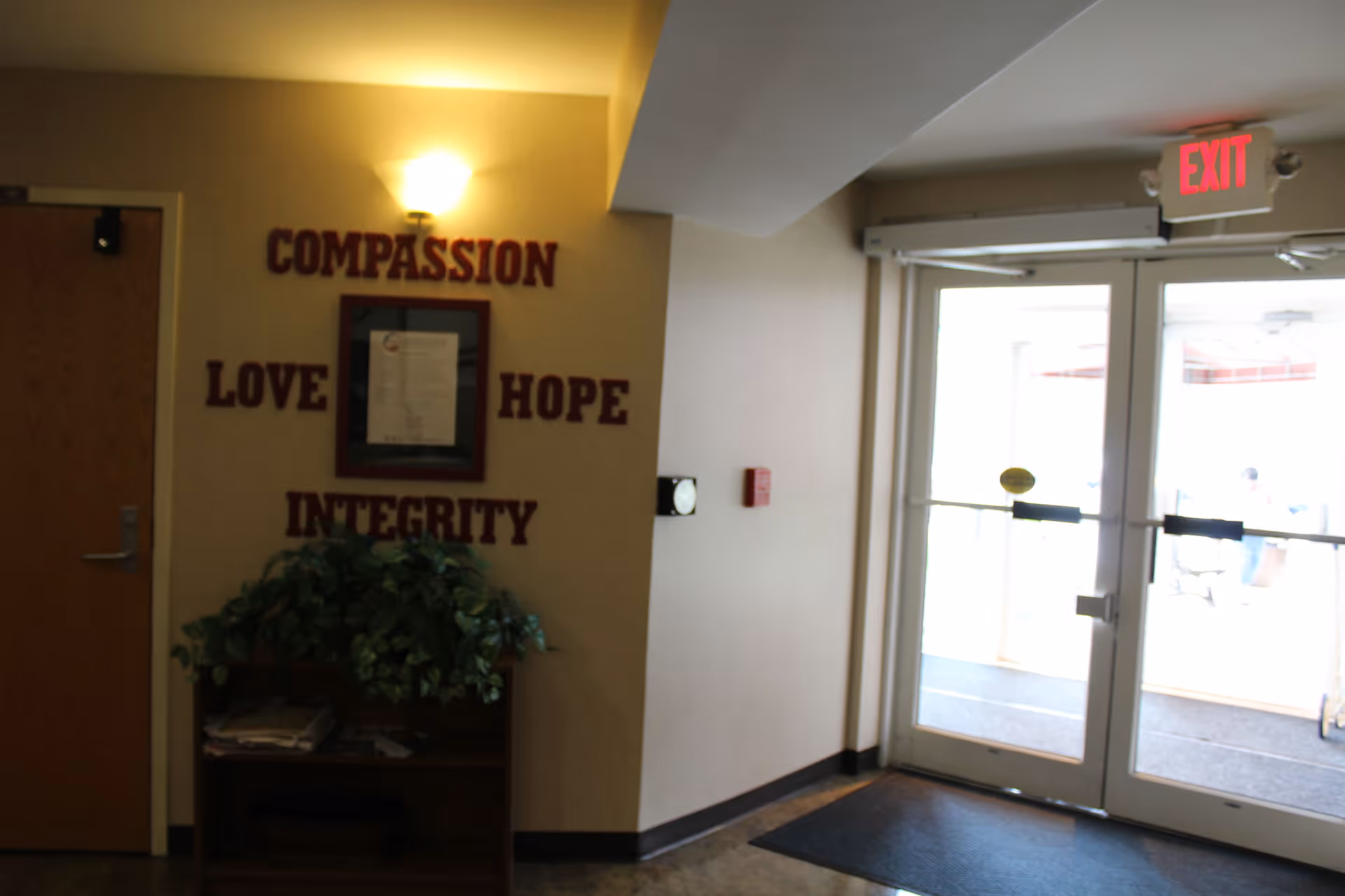 Interior view of an entrance area in St. Agnes Apartments featuring double glass exit doors with an illuminated red EXIT sign above. To the left, there is a wall with words compassion, love, hope, and integrity displayed around a framed document. Below the words is a small wooden shelf with a green leafy plant and some papers.