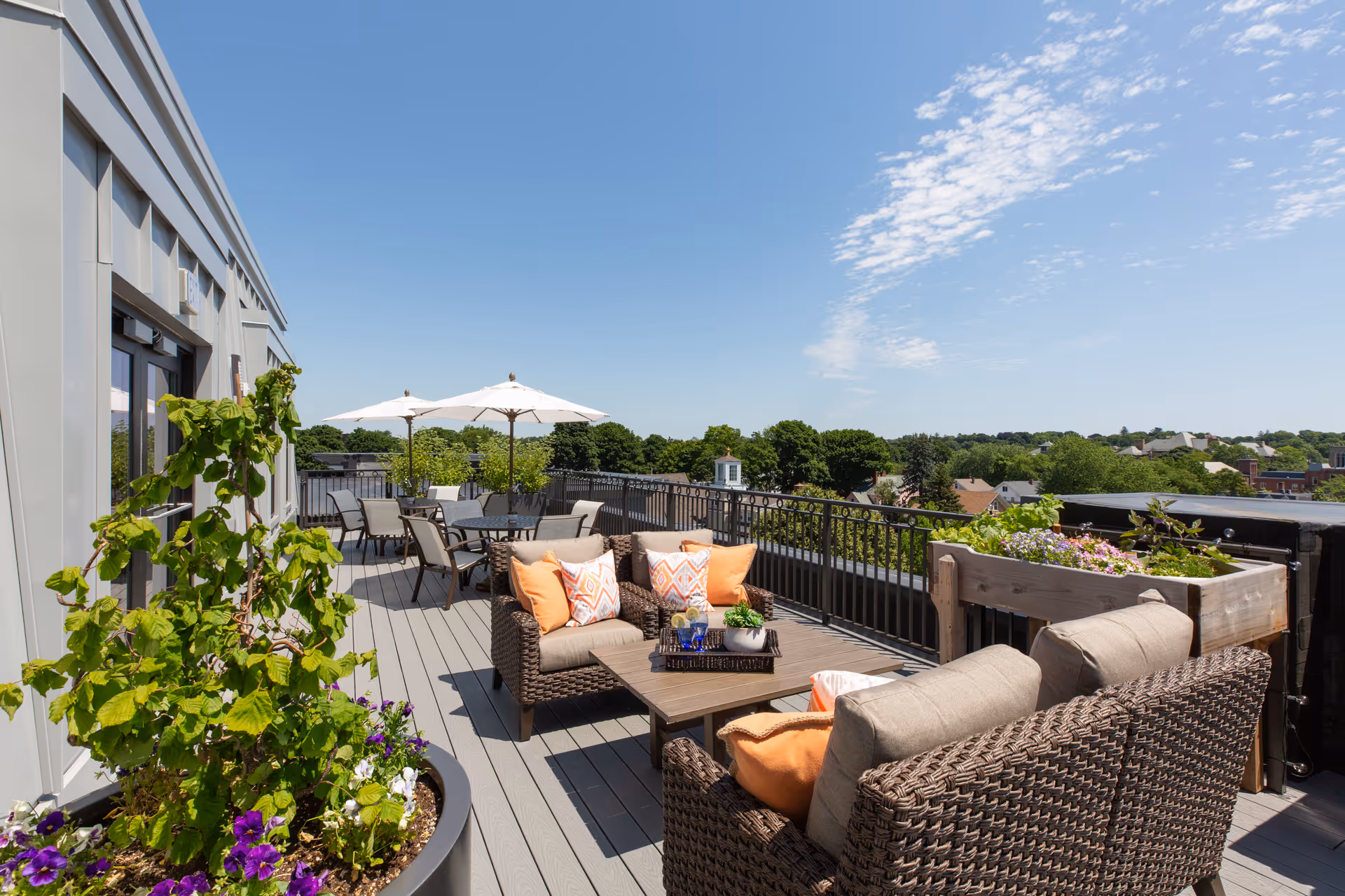 Outdoor patio area with wicker furniture including cushioned chairs and a coffee table, decorated with orange and patterned pillows. There are large potted plants and flower boxes, several tables with umbrellas, and a view of trees and rooftops under a clear blue sky.