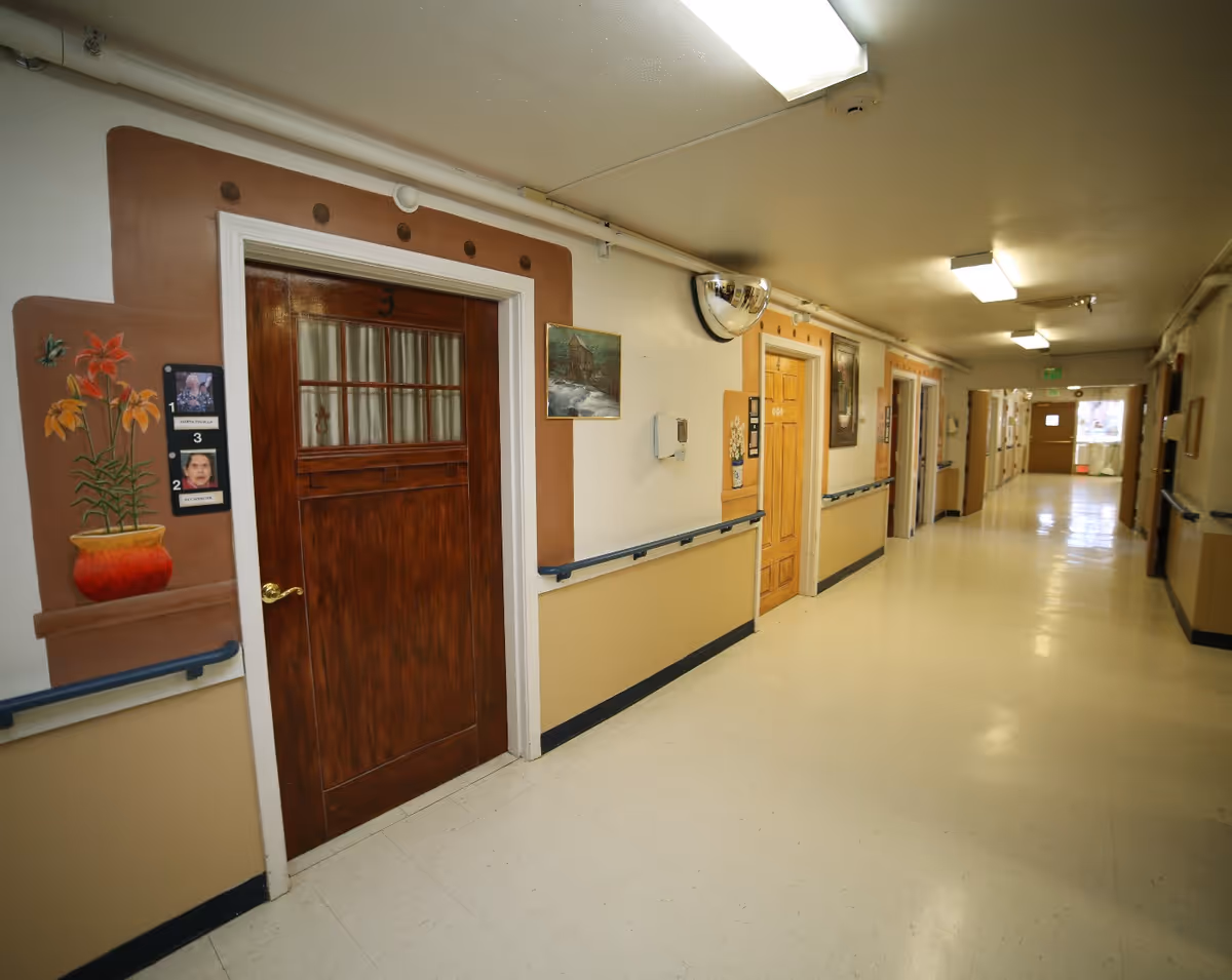 A long, well-lit hallway in a senior living facility with several wooden doors on the right side, each door decorated with painted frames and small paintings. The walls are painted beige and white with handrails along the length of the hallway. There is a convex security mirror mounted on the wall near the ceiling.