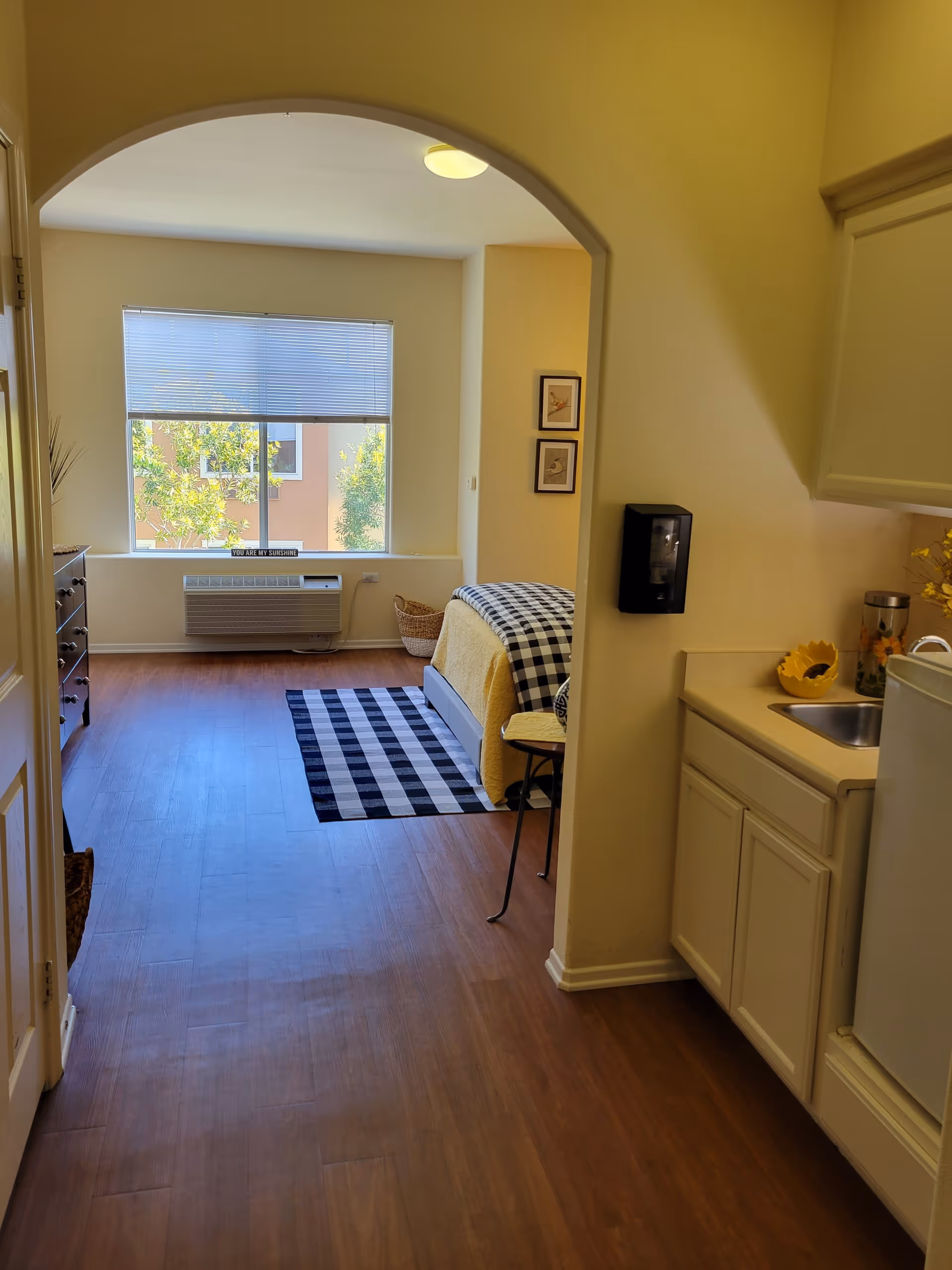 View of a senior living facility room at Cypress Place showing a small kitchenette area with a sink and mini fridge on the right, and a bedroom area with a bed covered in a yellow blanket and black-and-white checkered throw. There is a window with blinds partially open letting in natural light, a black-and-white checkered rug on the wooden floor, and some framed pictures on the wall.