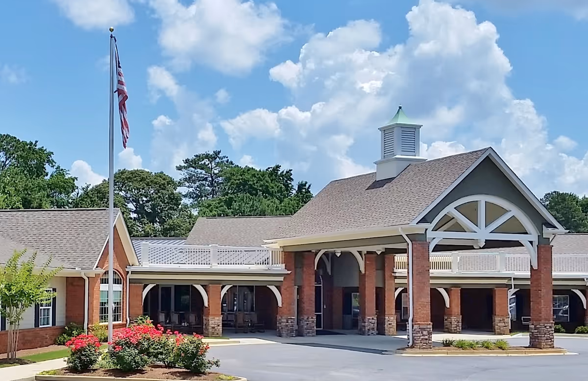 Front entrance of a red-brick senior living building with a covered porte-cochere, flagpole, and landscaped flower beds.