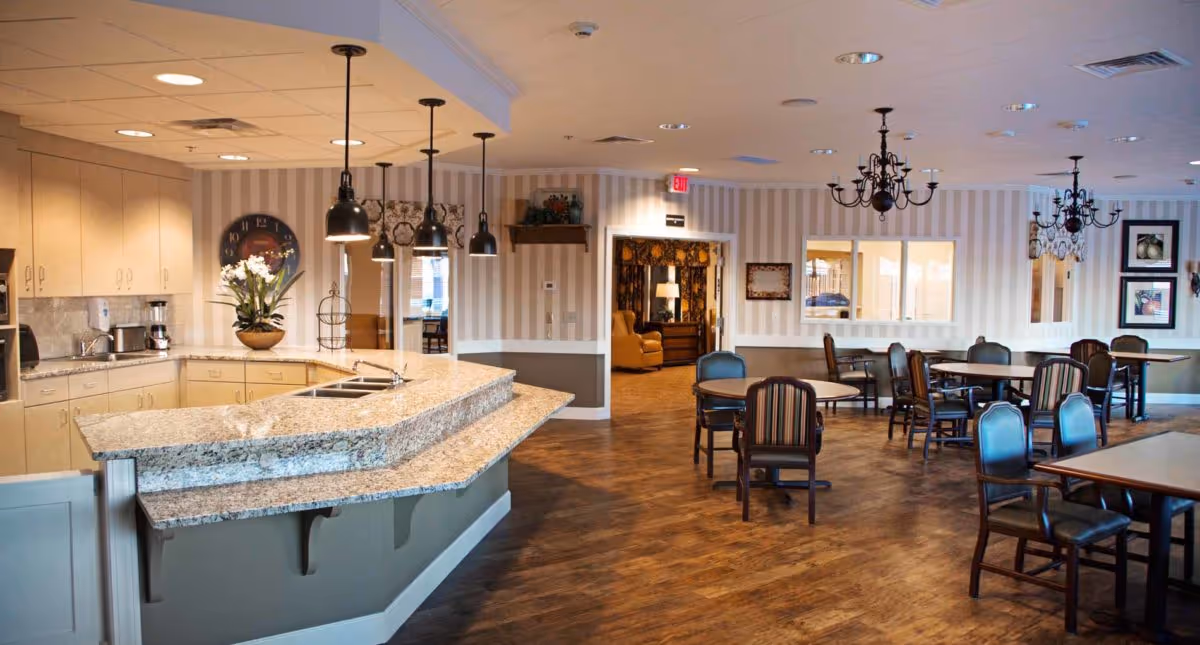 A spacious dining area with multiple round and rectangular tables surrounded by chairs. To the left, there is a kitchen counter with a granite countertop, a sink, and hanging pendant lights. The walls have striped wallpaper and there are chandeliers hanging from the ceiling. In the background, a cozy seating area with armchairs is visible through an open doorway.