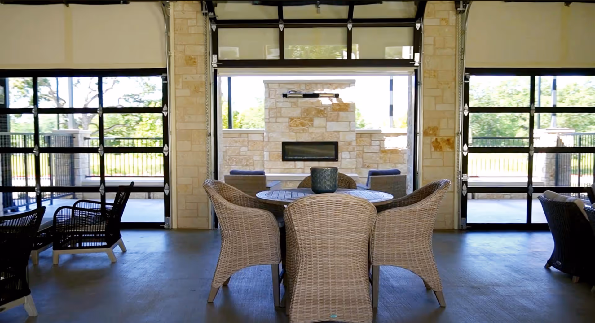 Indoor seating area with wicker chairs arranged around a round table, large glass garage-style doors open to an outdoor space with a stone fireplace and additional seating visible outside.