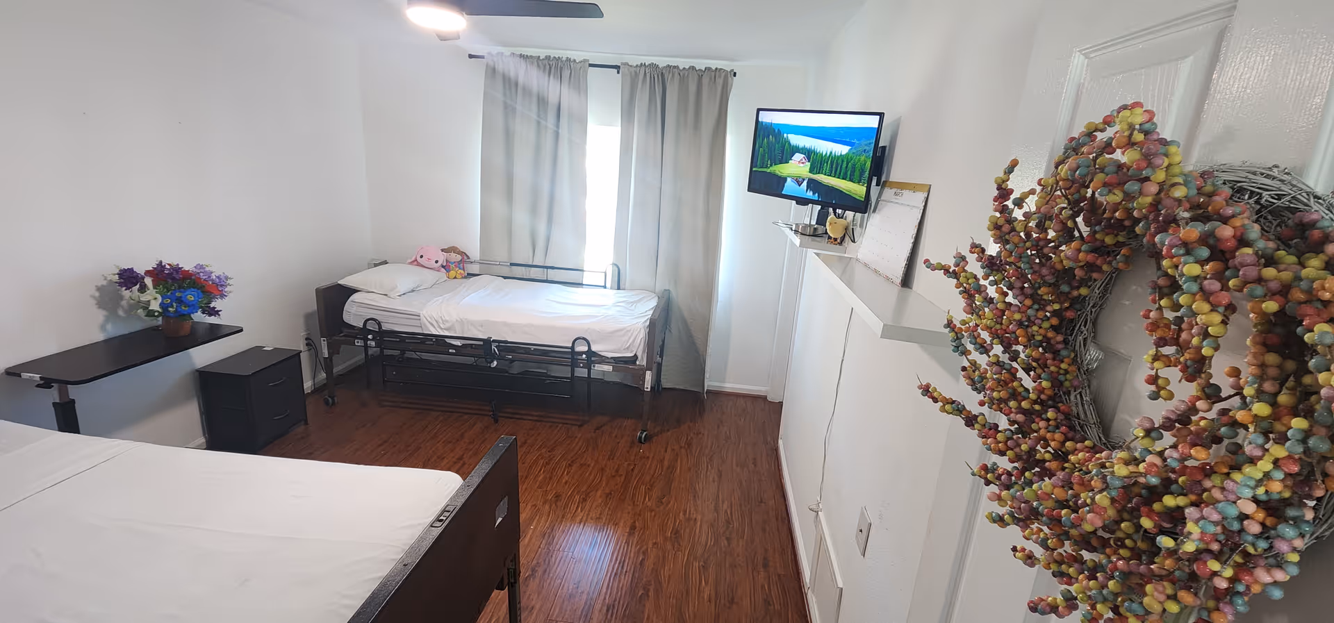 A simple, clean bedroom with two single beds, one against the left wall and the other near a window with gray curtains. There is a small black nightstand with a vase of colorful flowers next to the left bed. A flat-screen TV is mounted on the wall opposite the beds, and a decorative wreath made of multicolored berries hangs on the right wall. The floor is wooden, and the room is well-lit with a ceiling fan light.
