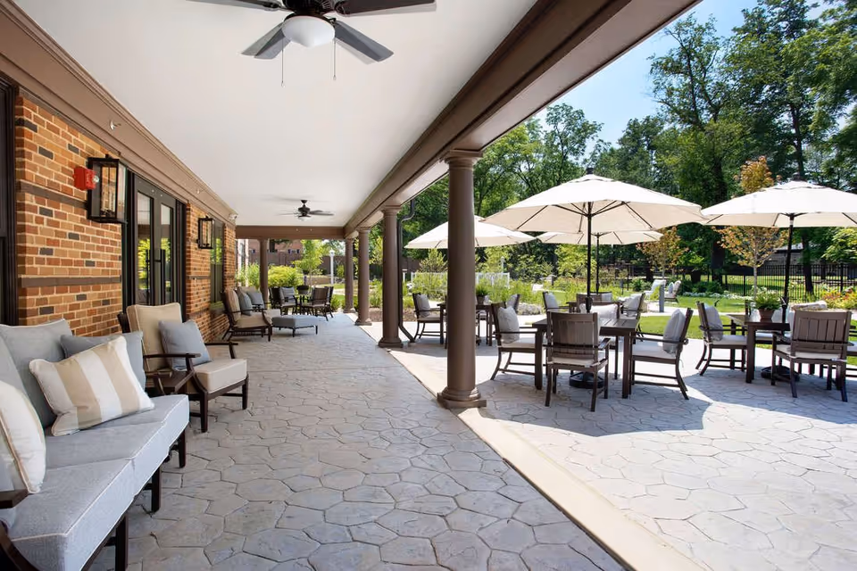 Covered outdoor patio area with cushioned seating along a brick wall on the left and several tables with chairs and large white umbrellas on the right, surrounded by greenery and trees under a clear sky.