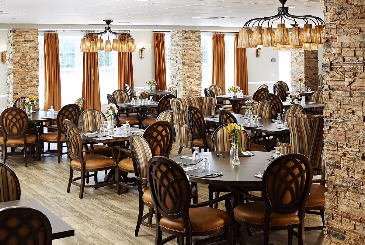 A bright and elegant dining room with multiple round and rectangular tables set with glasses, napkins, and silverware. The room features wooden floors, stone pillars, large windows with orange curtains, and decorative chandeliers hanging from the ceiling. Each table has a small vase with fresh flowers.