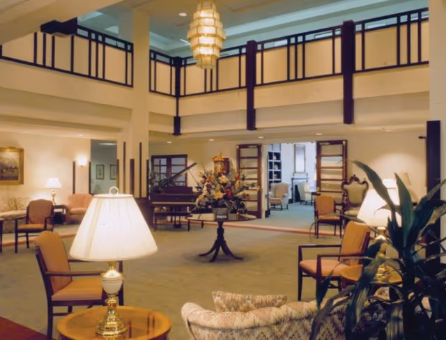 Spacious and well-lit interior common area with a high ceiling and balcony railing above. The room features multiple seating arrangements with cushioned chairs and sofas, several table lamps, a central round table with a large floral arrangement, and a grand piano in the background. The walls are light-colored, and there are open double doors leading to another room with additional seating.