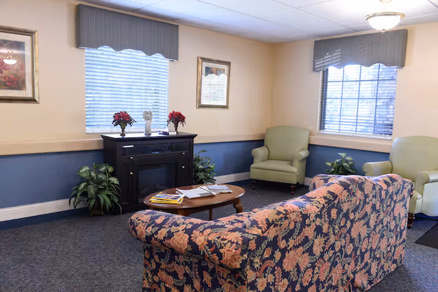 Cozy sitting room with a floral sofa, two green armchairs, a coffee table, and a fireplace console beneath two windows.