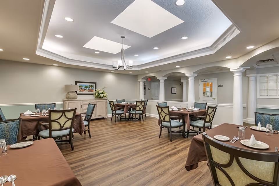 A dining room in a senior living facility with several round tables covered with brown tablecloths, each set with plates, napkins, glasses, and silverware. The room features wooden flooring, white columns, a sideboard with a lamp and flower arrangement, and a ceiling with recessed lighting and a chandelier.