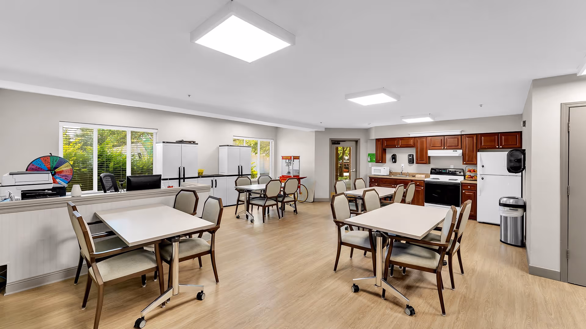 A bright and spacious common area with several tables and chairs arranged for dining or activities. The room features large windows letting in natural light, a kitchen area with wooden cabinets, a stove, microwave, and refrigerator. There is also a counter with office equipment and a colorful prize wheel on the left side.