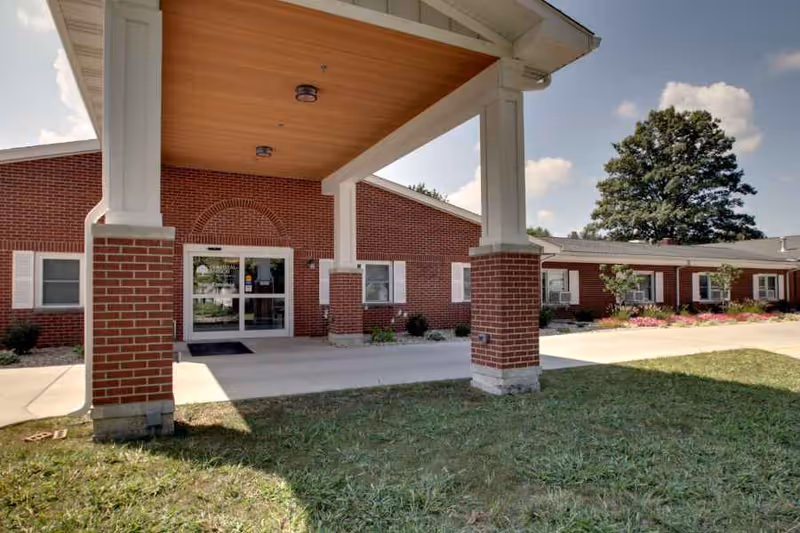 Exterior view of a single-story brick building with a covered entrance supported by large white columns. There is a concrete walkway leading to glass double doors. The building has white window shutters and landscaping with grass, bushes, and flowers. A large tree is visible in the background under a partly cloudy sky.