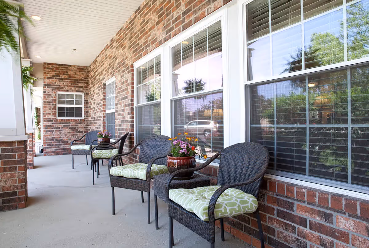 Covered brick-front porch with wicker chairs, green patterned cushions, and potted flowers in front of large windows.