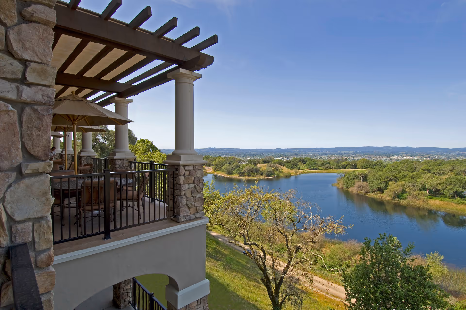 View from a stone and column-supported balcony with outdoor seating and umbrellas overlooking a river, trees, and distant hills under a clear blue sky.