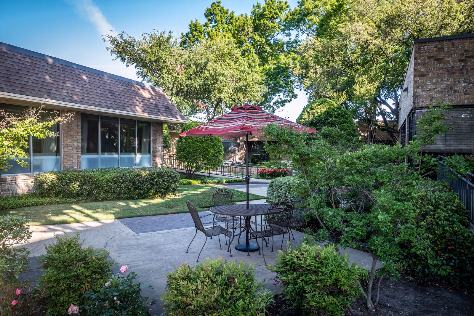 Outdoor patio area at Christian Care Nursing & Rehabilitation featuring a round metal table with four chairs and a red and white striped umbrella. The patio is surrounded by green bushes, trees, and well-maintained landscaping with a brick building in the background under a clear blue sky.