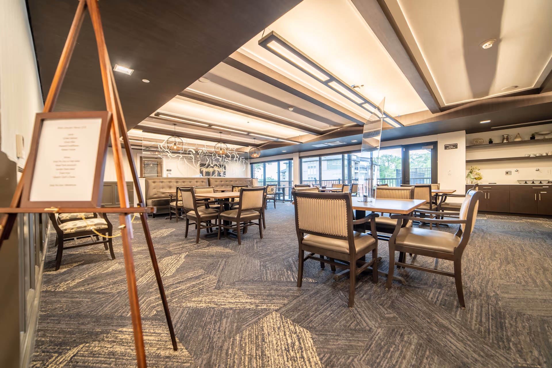 Open dining room with wooden tables and upholstered chairs, decorative ceiling lights, and large windows.