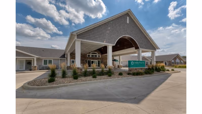Exterior view of Cedarhurst Senior Living of Springfield showing the main entrance with a covered driveway, landscaped plants, and a sign with the facility's name under a partly cloudy sky.