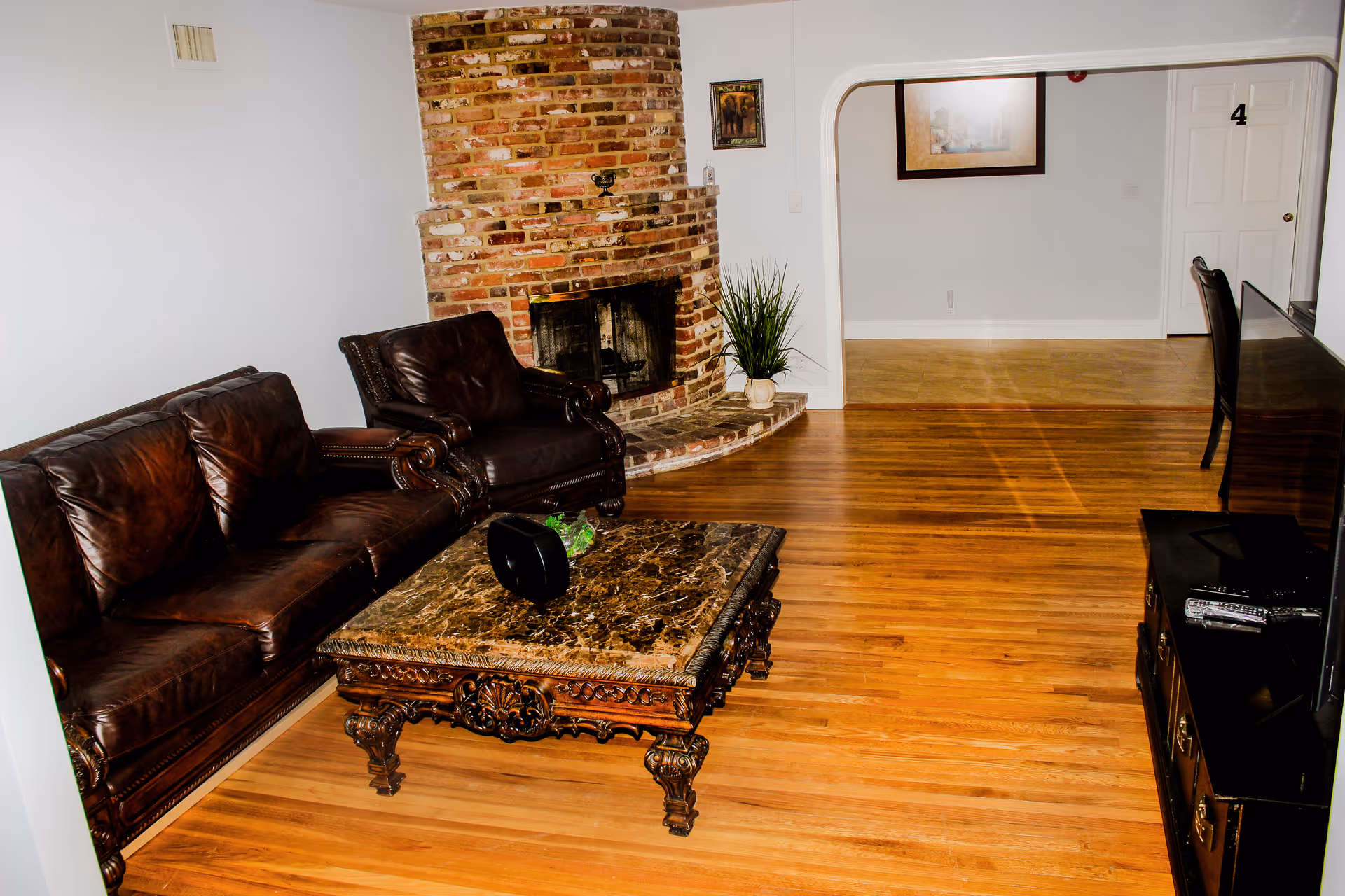 A living room with a brown leather sofa and armchair, a decorative wooden coffee table with a marble top, a brick fireplace in the corner, a potted plant next to the fireplace, and a flat-screen TV on a stand. The room has hardwood floors and white walls, with an open doorway leading to another room with a white door marked with the number 4.