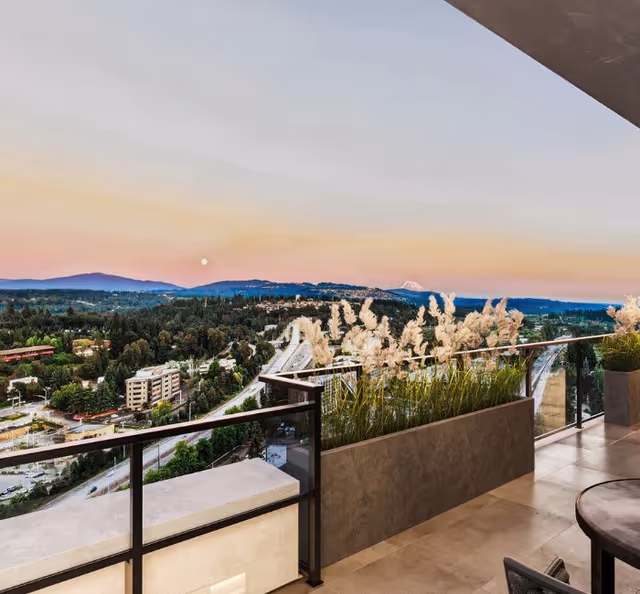 View from a balcony overlooking a scenic landscape with trees, buildings, and distant mountains under a pastel-colored sky at sunset. The balcony has a glass railing with a planter box containing tall ornamental grasses.