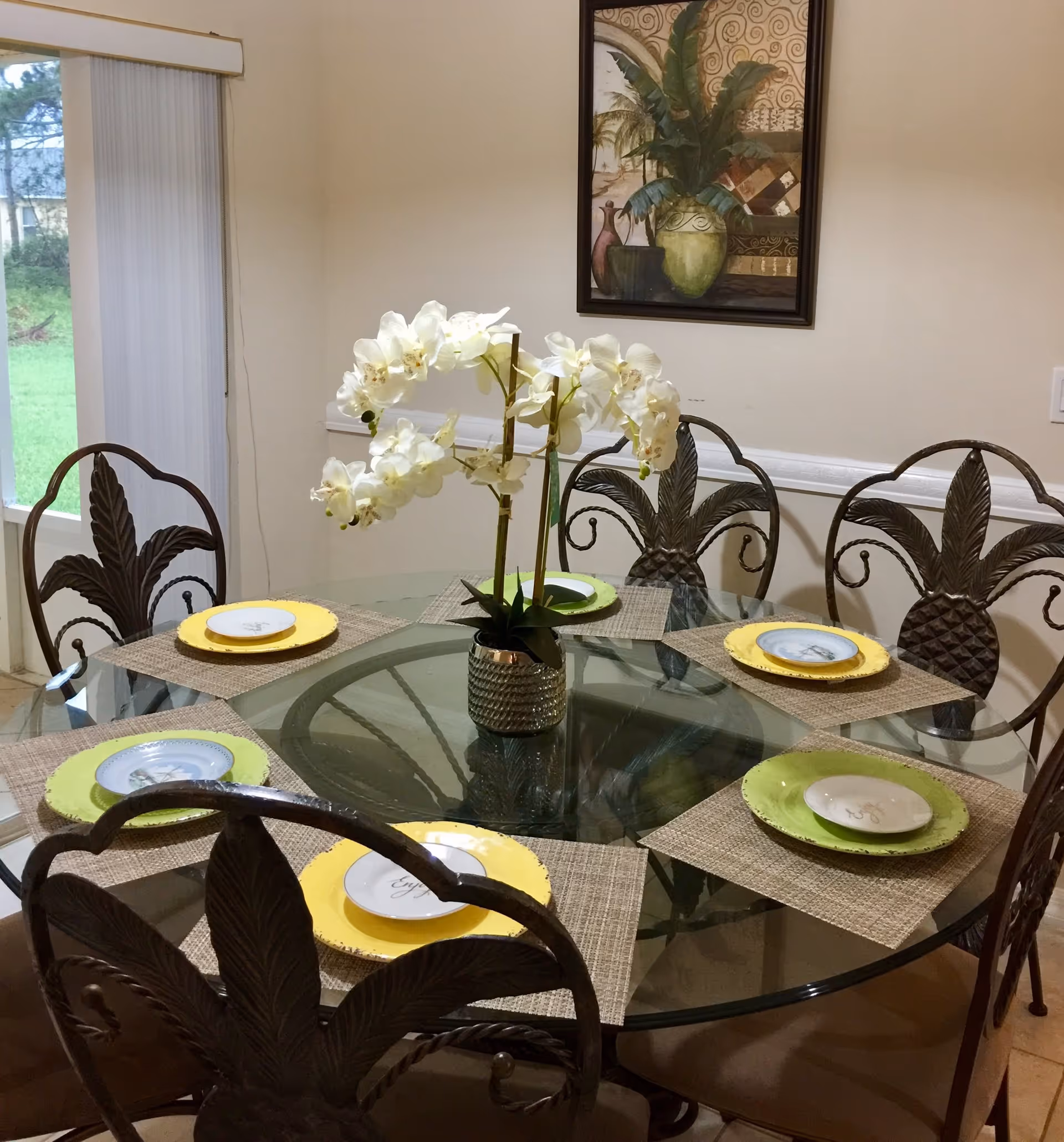 A dining room with a round glass table set for six people. The table has woven placemats, yellow and green charger plates, and small white plates on top. A potted white orchid is placed in the center of the table. The chairs have decorative metal backs with a leaf design. A window with vertical blinds is on the left, and a framed painting of plants hangs on the beige wall.