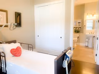 A bright assisted living bedroom with a hospital-style bed featuring a red heart-shaped pillow. The room has white double closet doors and an open doorway leading to a bathroom with a sink and mirror visible.