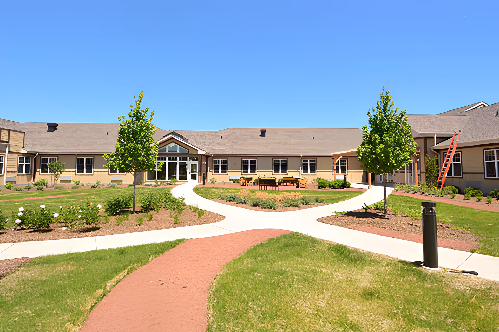 Exterior view of a single-story memory care community building with beige siding and a brown roof, surrounded by a landscaped garden with green grass, small trees, and flower beds under a clear blue sky. There are paved walkways leading to the entrance and outdoor seating areas with benches.