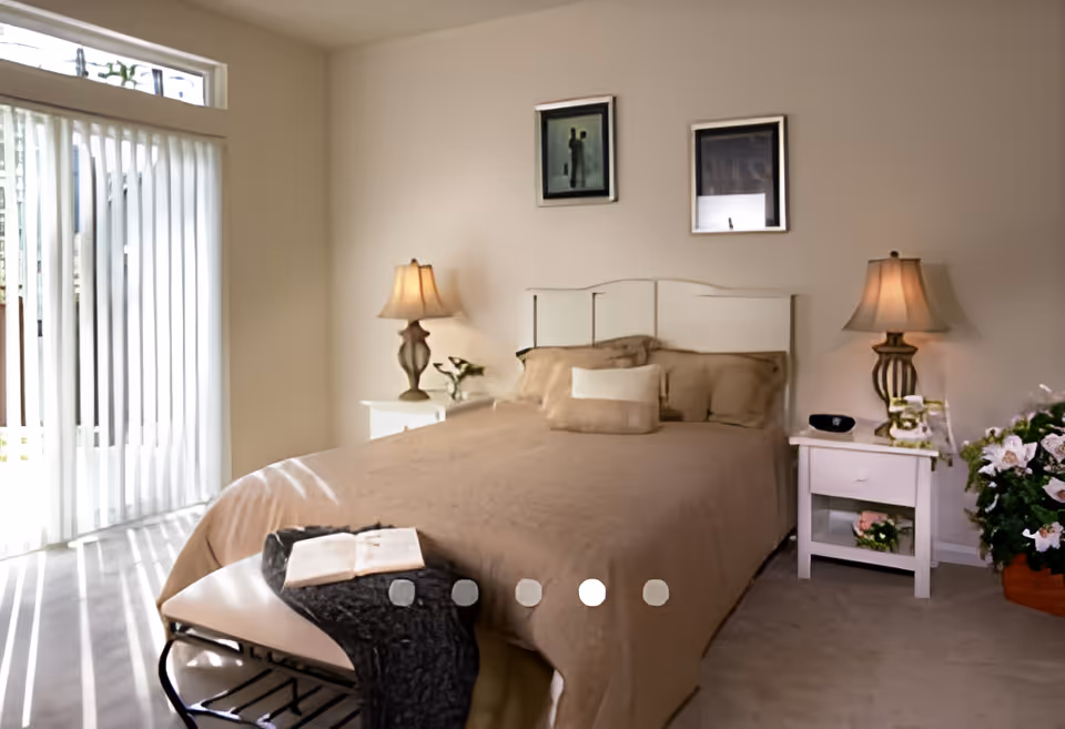 Well-lit bedroom with a made bed, two nightstands and lamps, sliding vertical-blind doors, and decorative artwork and plants.