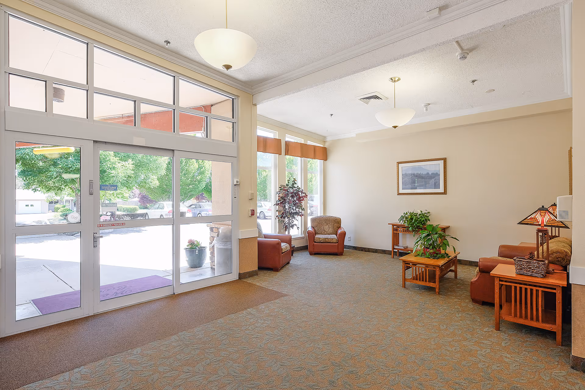 Bright reception lobby with glass entrance doors, upholstered chairs, wooden tables, plants, and pendant lights.