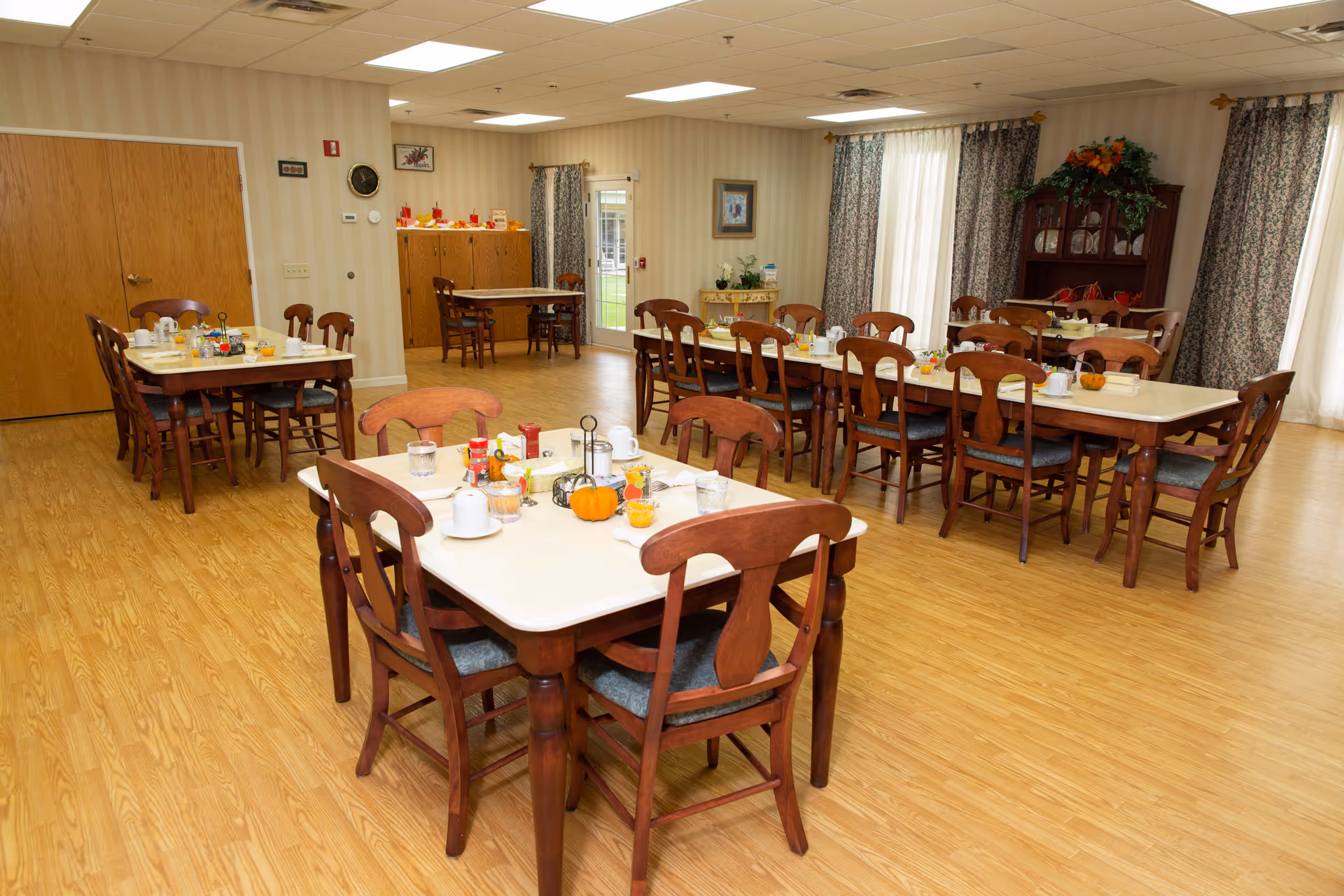 A dining room with several wooden tables and chairs arranged neatly. Each table is set with cups, glasses, condiments, and small decorative pumpkins. The room has wood flooring, beige striped wallpaper, and large windows with patterned curtains allowing natural light to enter. There is a wooden cabinet with glass doors and a floral arrangement on top in the background.