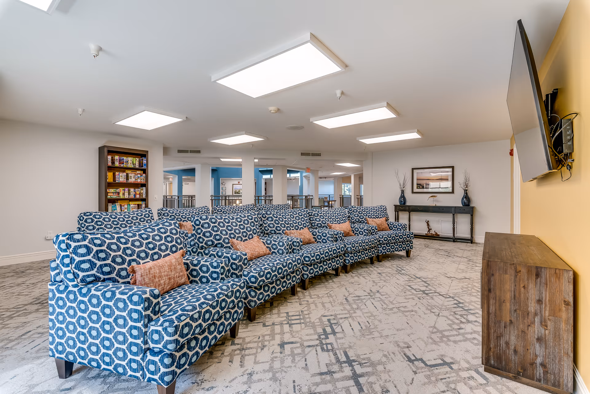 A bright senior living common room with patterned blue armchairs arranged in rows facing a wall-mounted TV and a console.