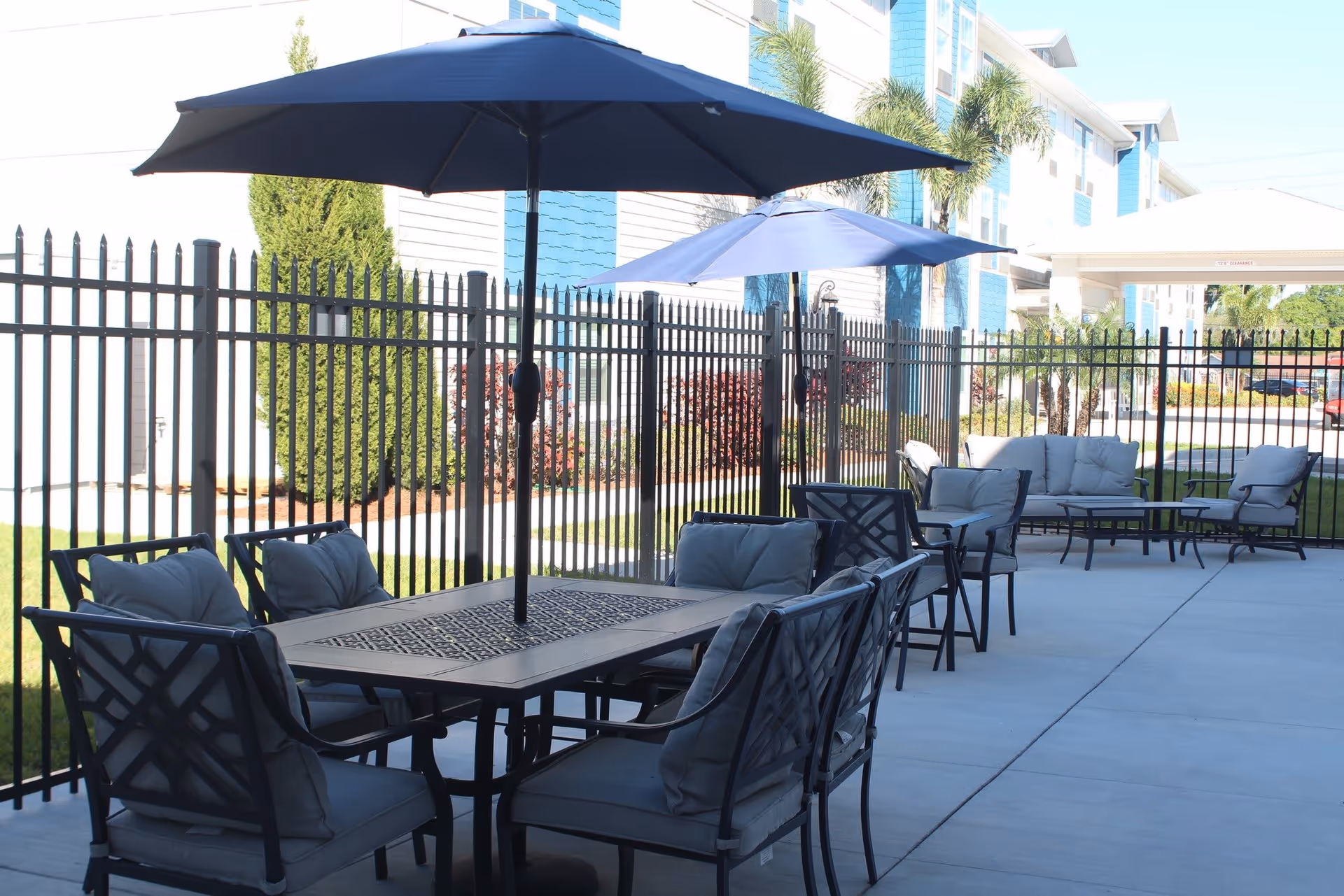 Outdoor patio area with cushioned chairs and tables shaded by large umbrellas, enclosed by a black metal fence with a building and greenery in the background.