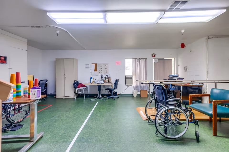 Physical therapy/activity room with wheelchairs, parallel bars, desks and chairs on a green floor.
