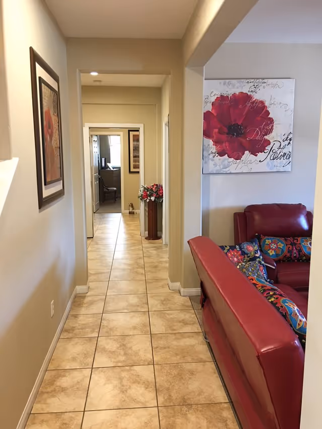 A tiled hallway in a senior living facility with beige walls and a framed picture on the left wall. At the end of the hallway is a room with a chair and a window. To the right of the hallway is a living room area with red leather furniture and colorful patterned pillows. A large painting of a red flower hangs on the wall above the furniture. A tall vase with flowers is placed near the doorway at the end of the hallway.