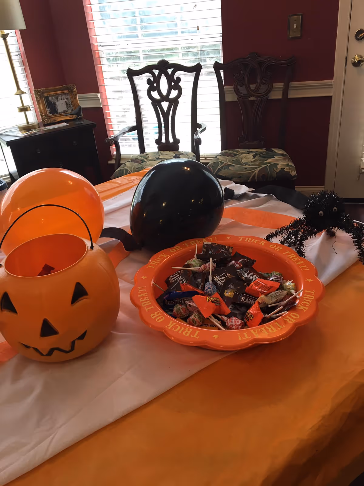 A dining table decorated for Halloween with a jack-o'-lantern candy bucket, an orange 'Trick or Treat' bowl full of candy, black balloons and a decorative spider, with chairs and a window in the background.