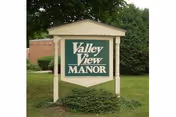 A large green and white sign with the text 'Valley View Manor' mounted on two white posts, situated on a grassy area with bushes and trees in the background.