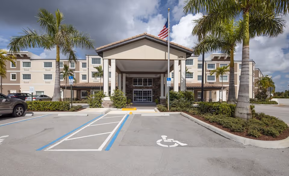 Front exterior view of a multi-story building with a covered entrance, palm trees, an American flag on a flagpole, and handicapped parking spaces in the foreground under a partly cloudy sky.