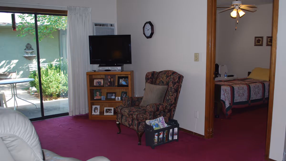 Interior view of a living room with a floral patterned armchair, a wooden shelf holding a TV and framed photos, and a sliding glass door leading to an outdoor patio. Adjacent to the living room is a bedroom with a bed covered in a quilt and a ceiling fan with lights.