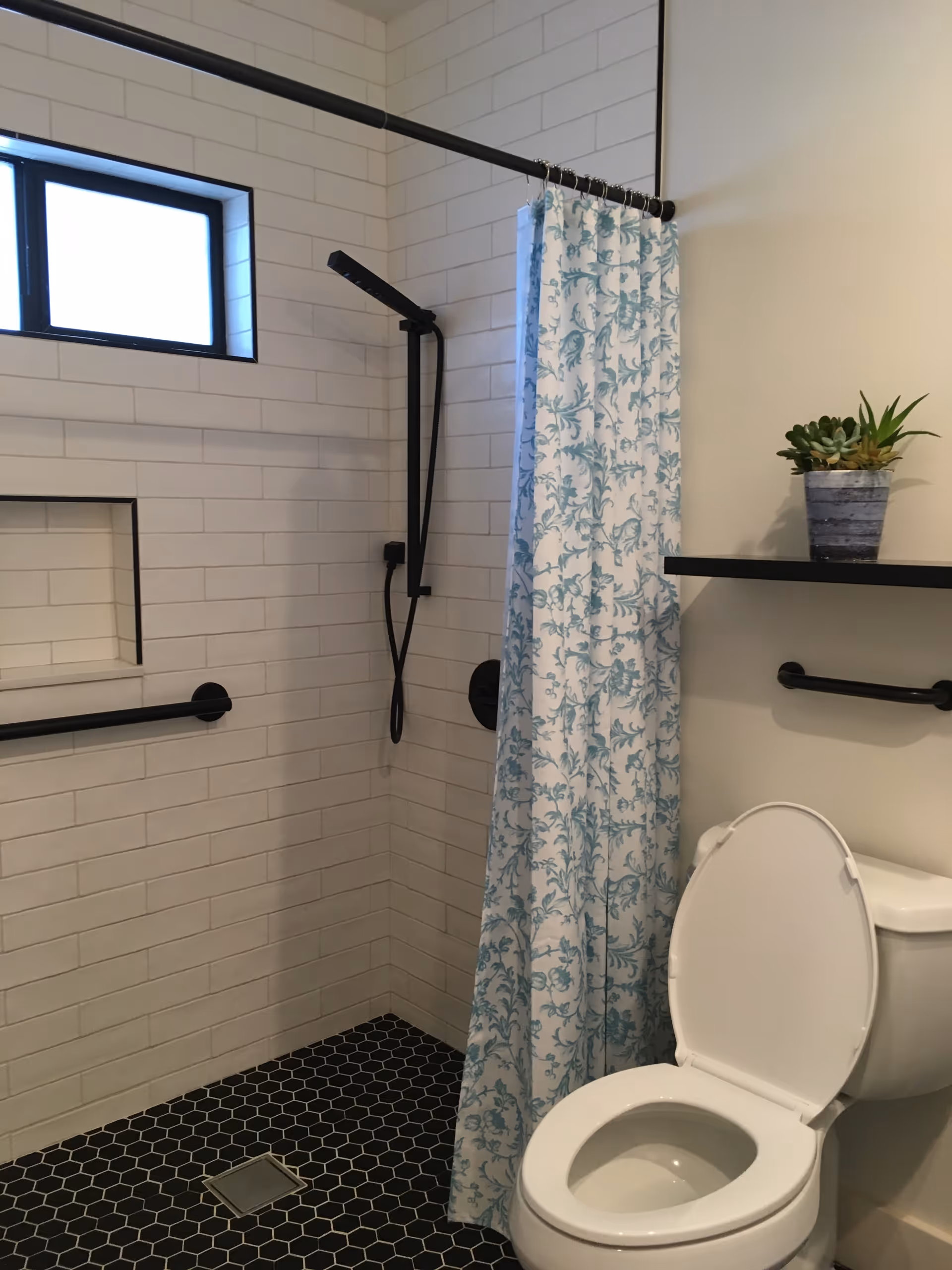 A modern bathroom featuring a walk-in shower with white subway tiles and a black hexagonal tile floor. The shower has a black showerhead and grab bar, and a blue and white floral shower curtain. There is a small window above the shower. Next to the shower is a white toilet with the lid open, and above it is a black shelf holding a small potted plant.